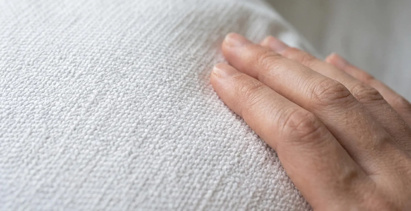 Gros plan macro sur le tissage serré d'une enveloppe de couette en percale de coton blanc révélant la densité des fils, main féminine effleurant délicatement le tissu, lumière rasante accentuant la texture et la qualité du textile